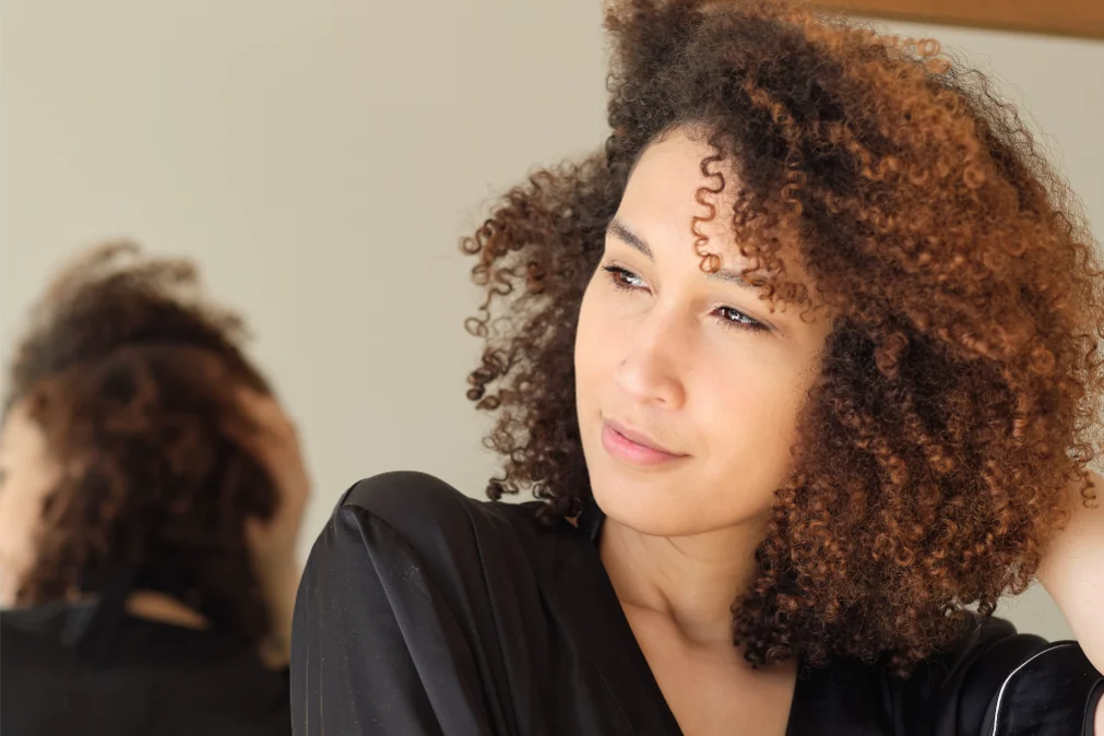 A woman with brown curly hair and a black shirt is looking in the distance in front of a mirror.