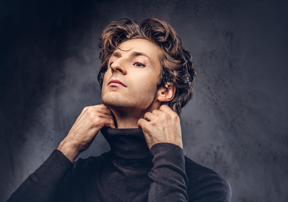 A man with medium length layered wavy brown hair wears a dark gray turtleneck and looks upwards, against a dark gray background.