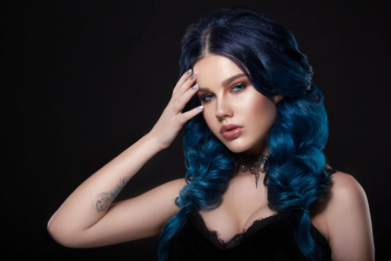 A woman with dark roots transitioning to bright blue curly hair, wearing a black lace top and choker, hand touching the forehead.