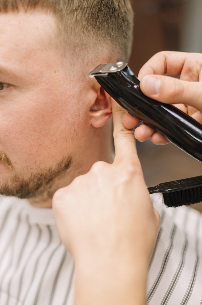 Barber using clippers to trim around a man's ear during a precise haircut, with striped cape visible.
