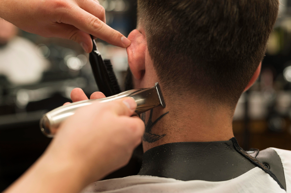 Barber trimming the neckline of a man's haircut with clippers, revealing a bird tattoo on the back of his neck.
