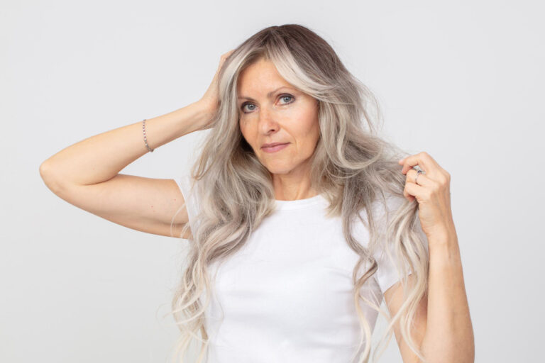 A woman in her 60s with long, blonde and gray hair wearing a white t-shirt and holding her hair up, with a serious expression.