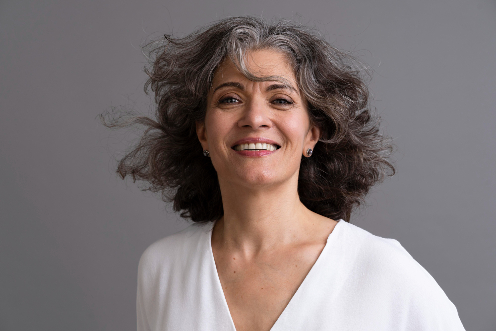 A smiling woman with gray, curly hair, wearing a white shirt stands against a gray background looking forward.