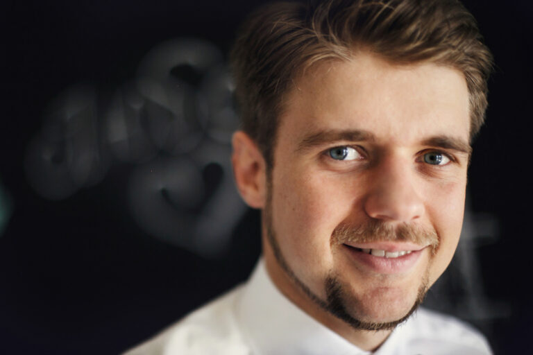 A smiling man with brown hair, blue eyes, and a goatee. He wears a white shirt.