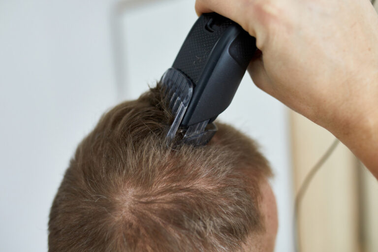 A hand uses a black electric hair clipper to cut the brown hair on top of a person's head.